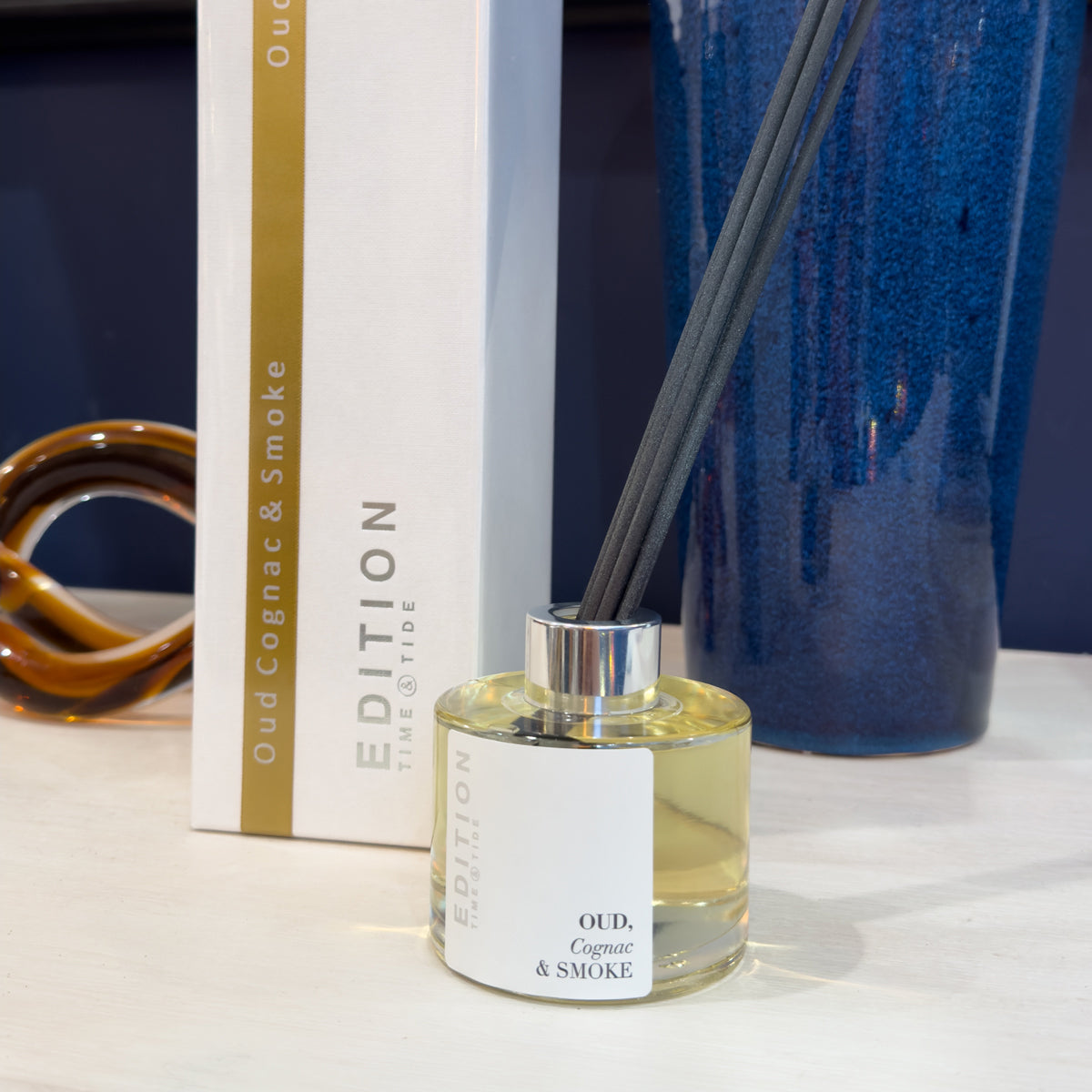 Diffuser bottle with 'Oud, Cognac & Smoke' fragrance on a white surface with a blue vase in the background.