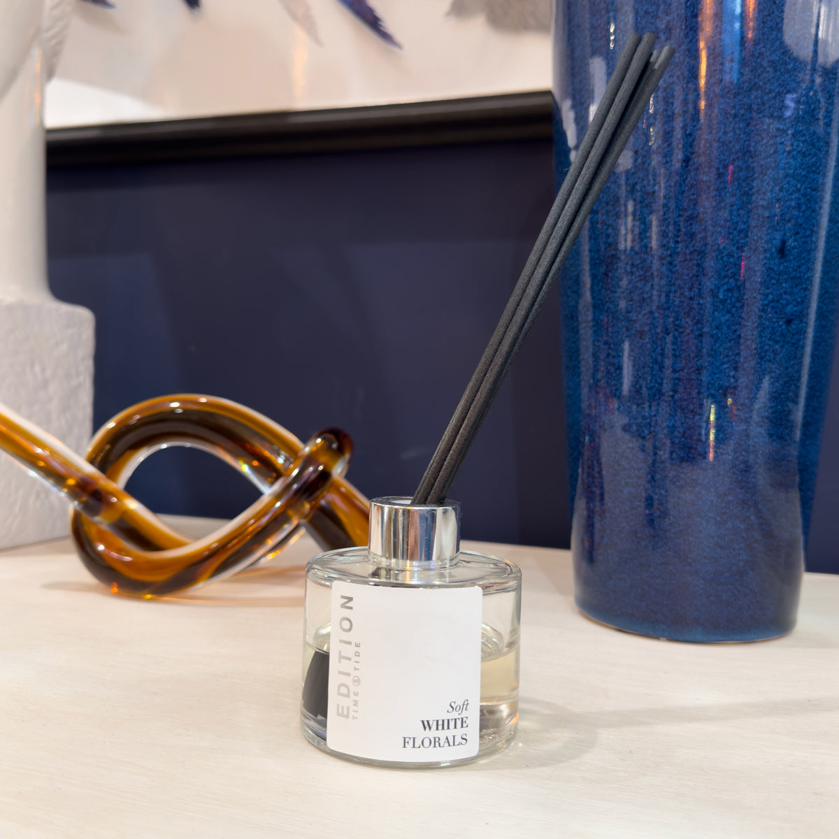Diffuser with reeds on a surface with a blue vase and decorative knot in the background