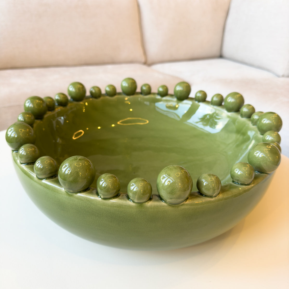 An olive green glossy ceramic bowl sitting on a white coffee table infront of a white sofa.