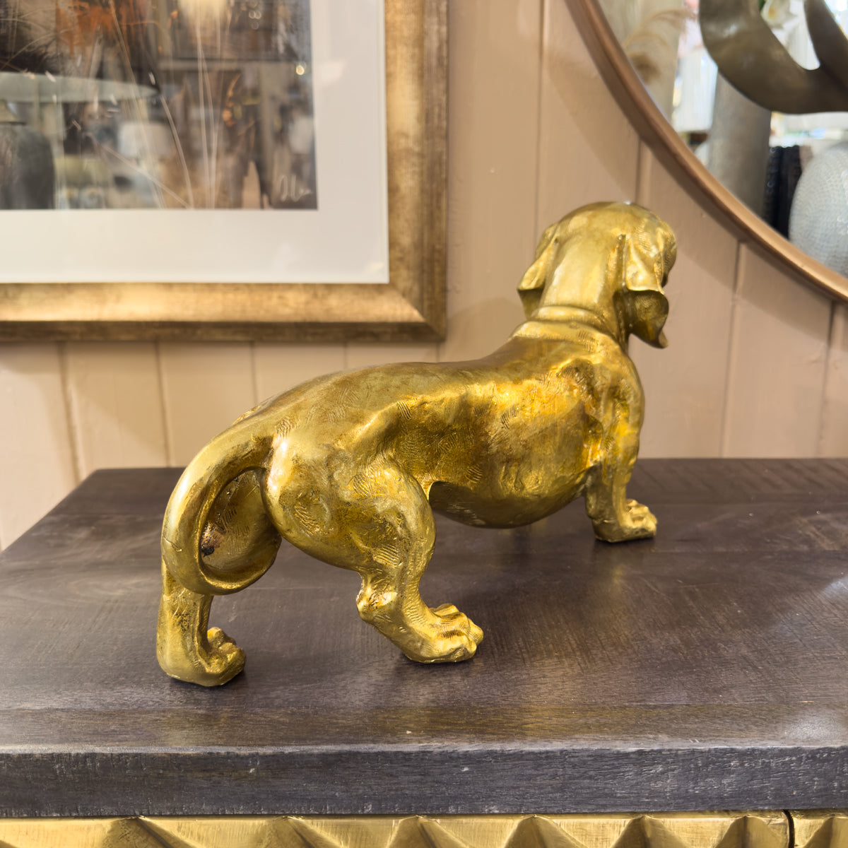 Gold dog statue on a dark surface with a mirror and framed picture in the background