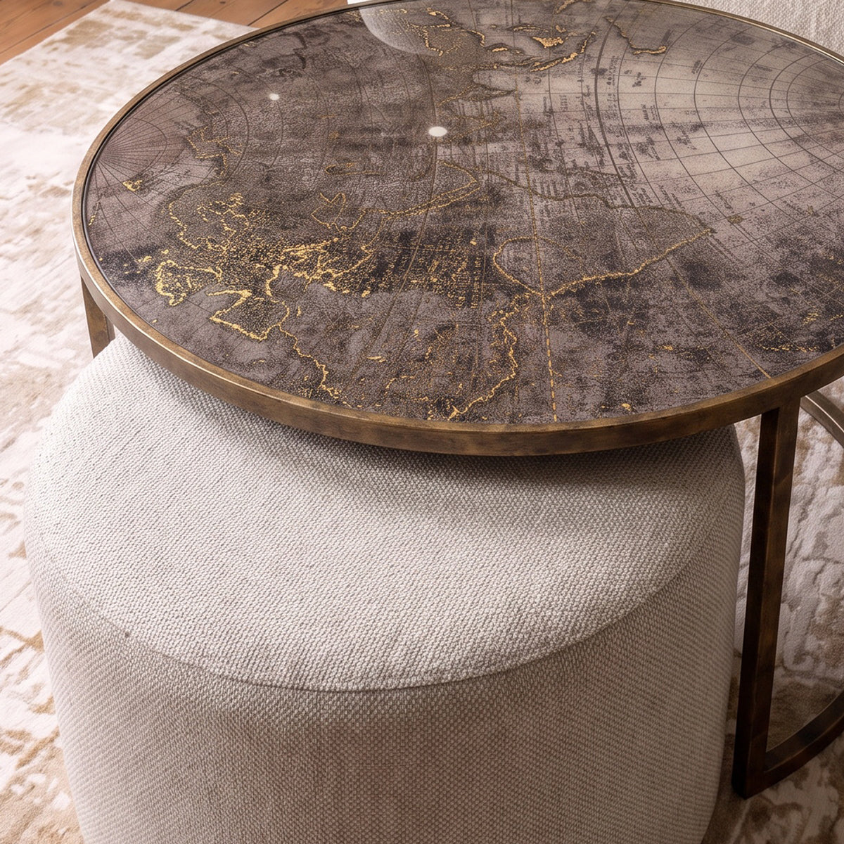 A round coffee table nest of 2with a distressed dark gray and gold world map print on the tabletop, supported by a cylindrical upholstered ottoman in light beige fabric.