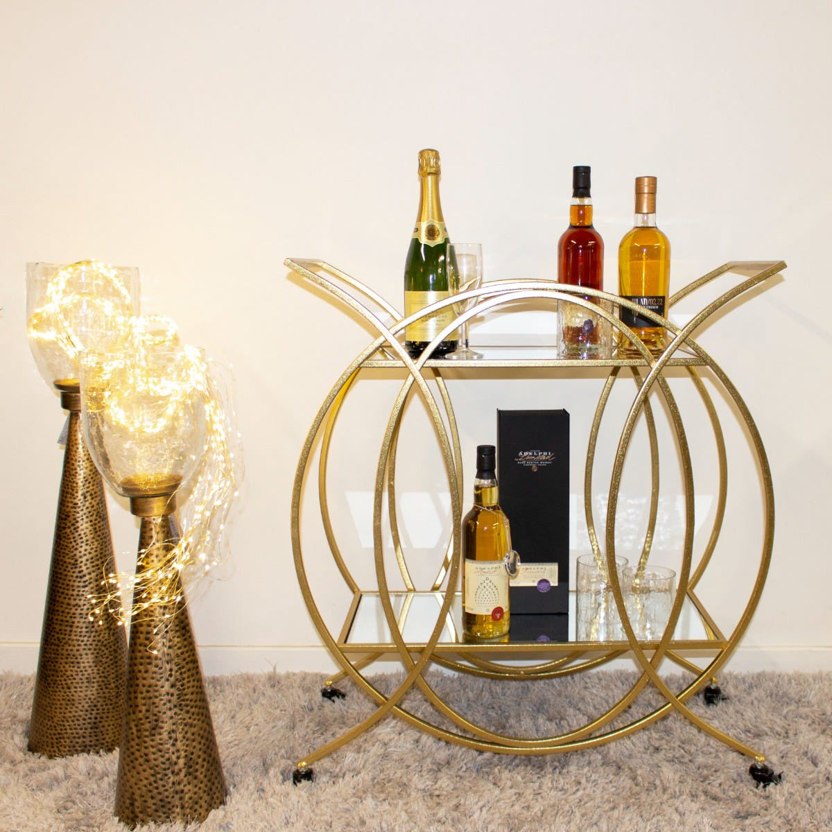 A golden deco style circular bar trolley with two mirrored shelves, displayed alongside a decorative lamp with a gold base.