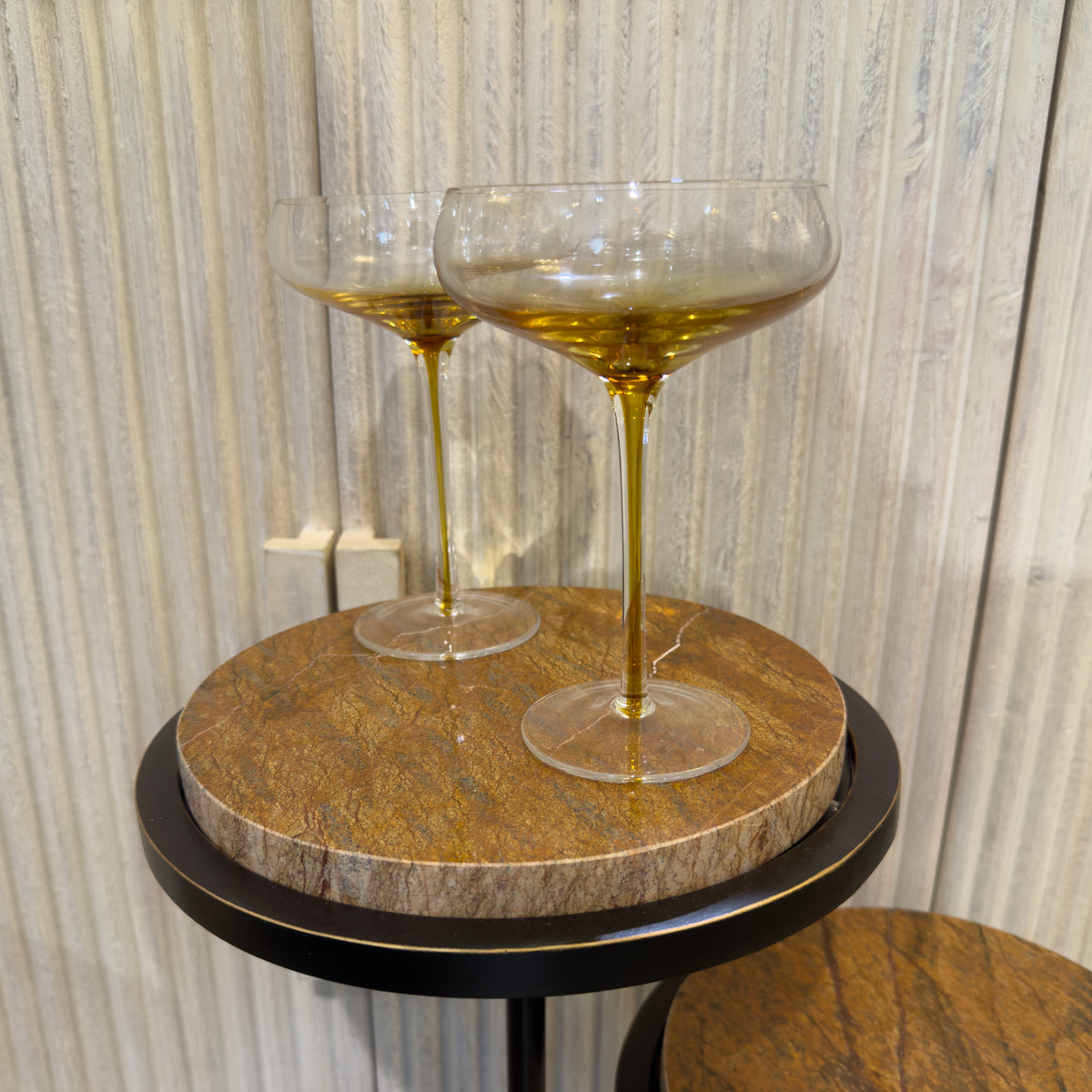 Two glass clear cocktail glasses with golden stems stand on a round marbled table against pale textured sideboard.