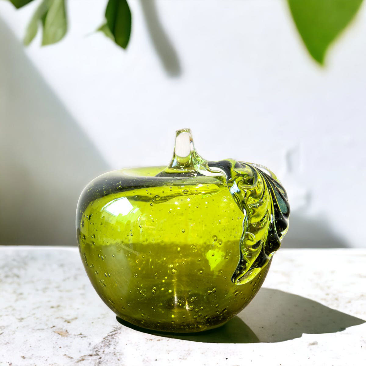 Glass apple-shaped object on a light surface with green leaves in the background