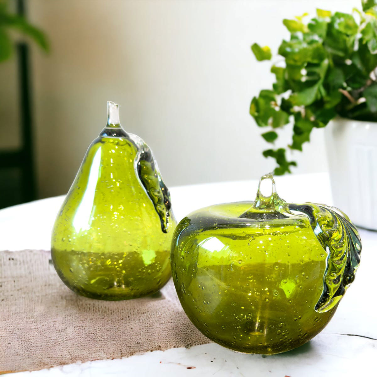 Two green glass fruit sculptures on a table with a blurred plant in the background