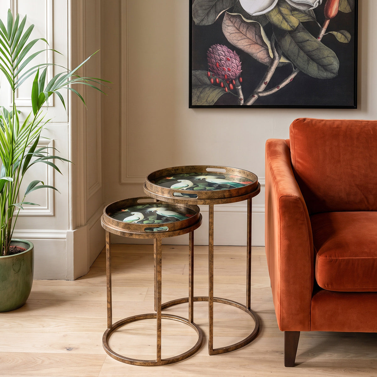 Two nested round nesting side tables with distressed bronze metal frames and inlay designs, styled in a living room next to an orange velvet sofa and a potted plant.