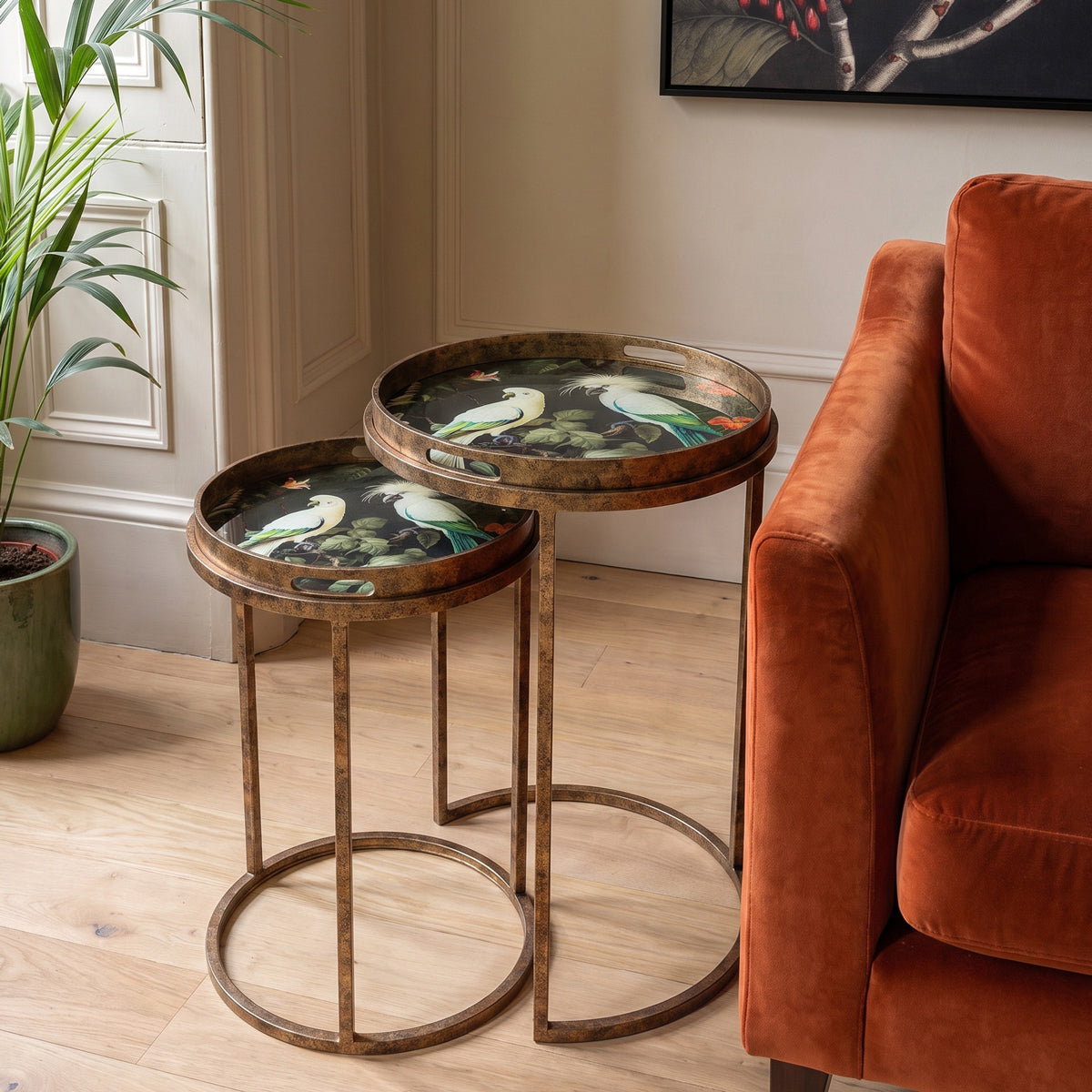 Two nested round nesting side tables with distressed bronze metal frames and round trays featuring a colorful bird and floral illustration, styled in a living room next to an orange sofa.