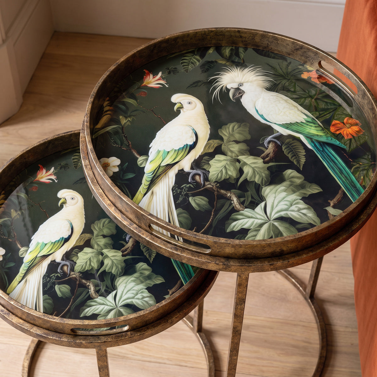 Two round nesting side tables with distressed bronze metal frames and legs, featuring a detailed tropical bird print with white cockatoos and green leaves on a dark background, set on a light wood floor.
