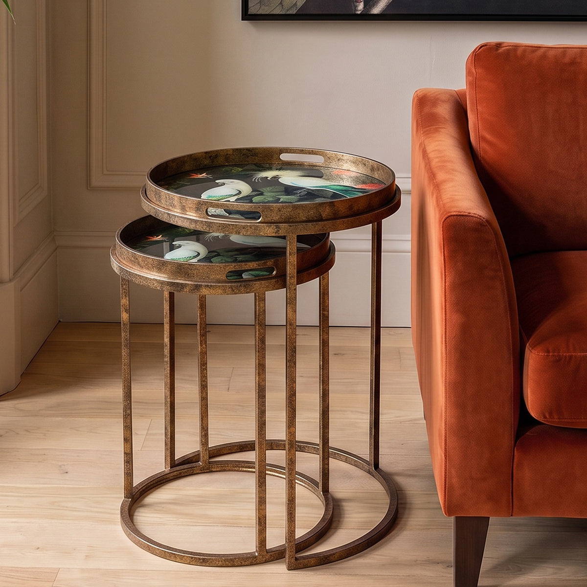 Two round nesting side tables with distressed bronze metal frames and oval trays featuring a colorful floral and bird pattern, styled in a living room next to an orange armchair.