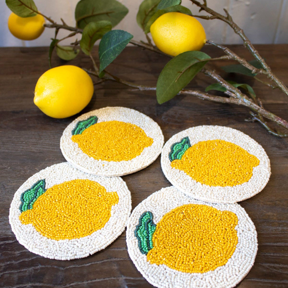 Four lemon-themed coasters on a wooden surface with lemons and branches in the background.
