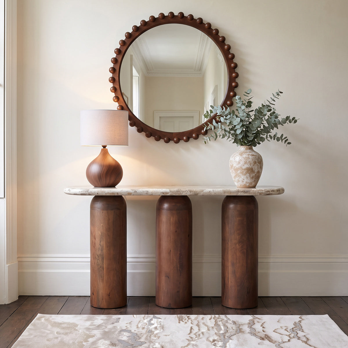 A styled interior scene showing a console table with three cylindrical dark wood legs and a light stone top, beneath a ball surround wooden mirror. 
