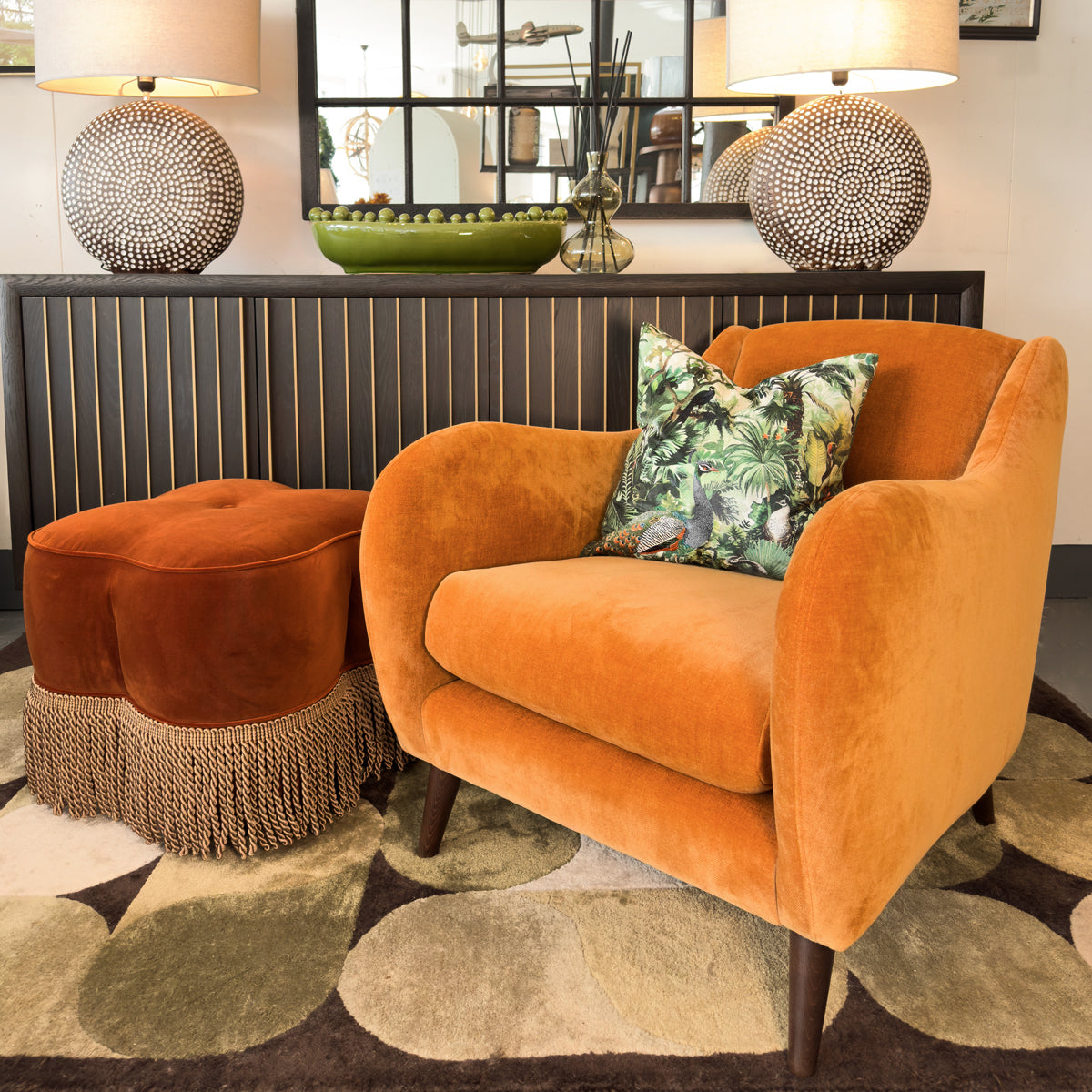 An orange velvet accent chair with walnut legs, positioned in a room with a footstool, decorative pillows, and a geometric rug.