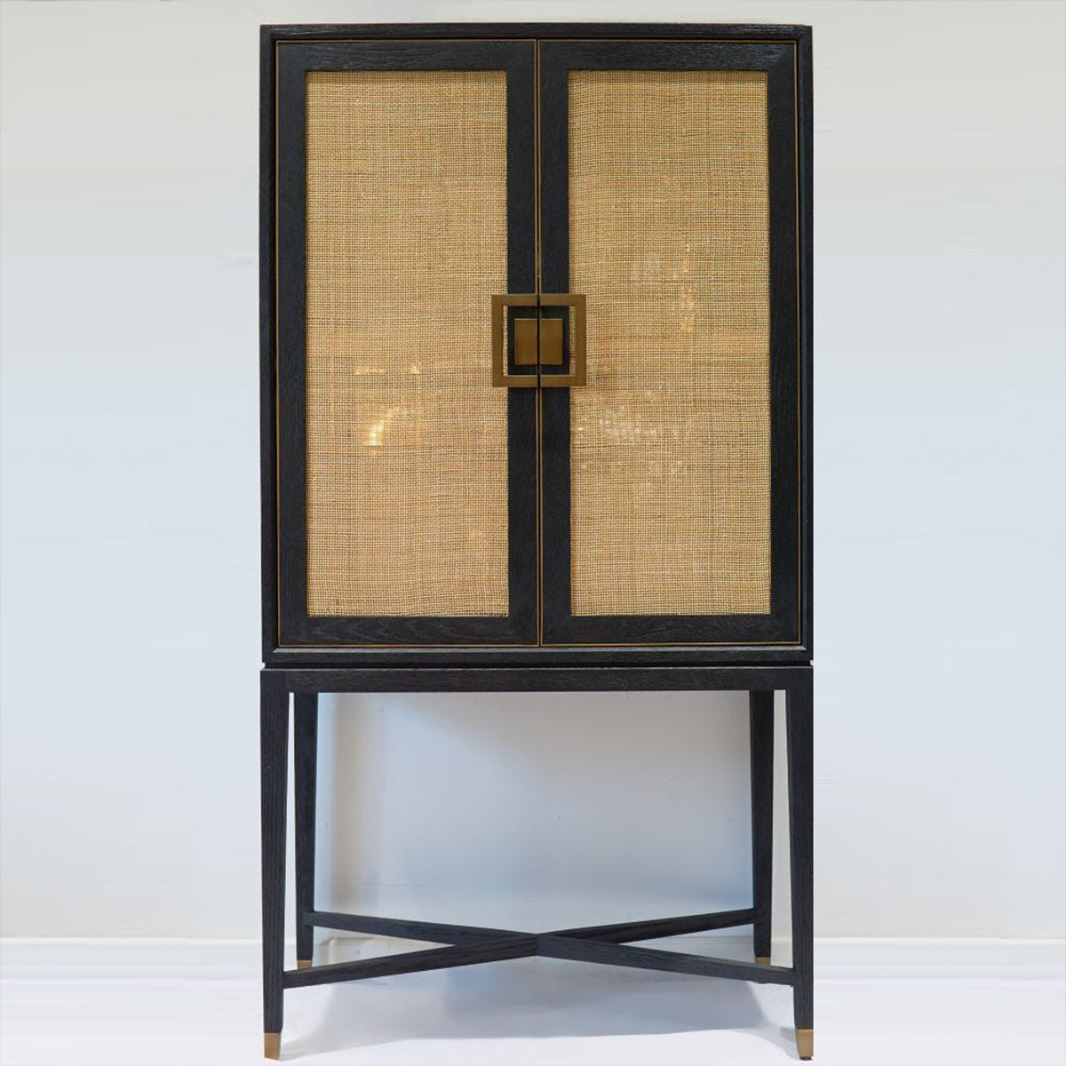 A black wood finish cabinet with natural rattan doors and brass handle details, standing on a cross-braced frame.