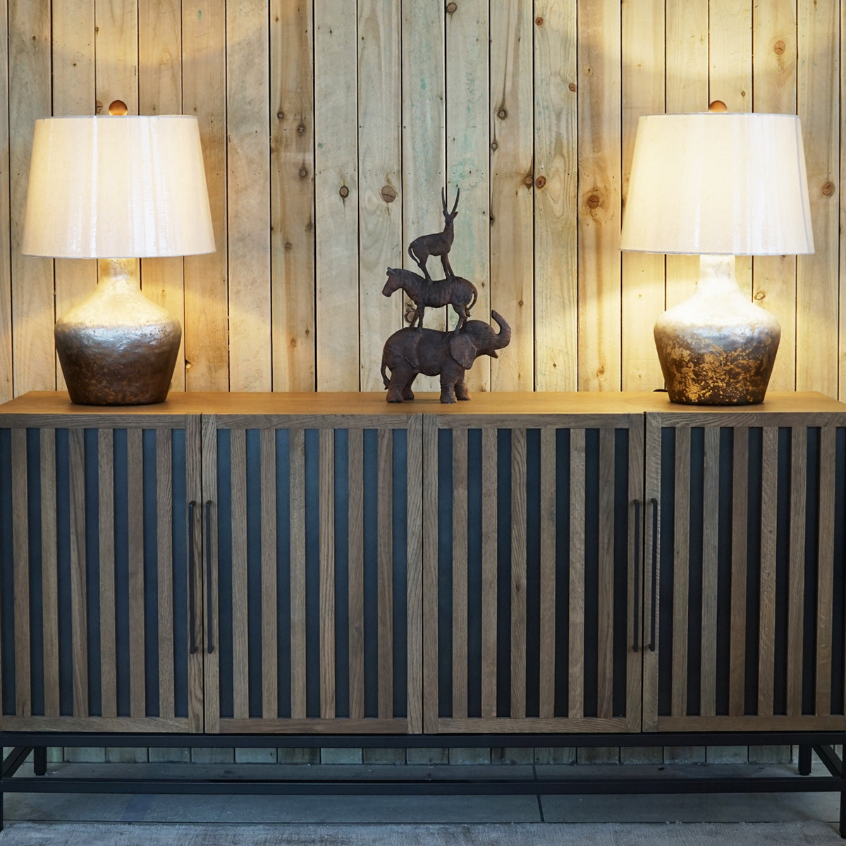 Wooden sideboard with two lamps and decorative animal sculpture against a wooden wall.