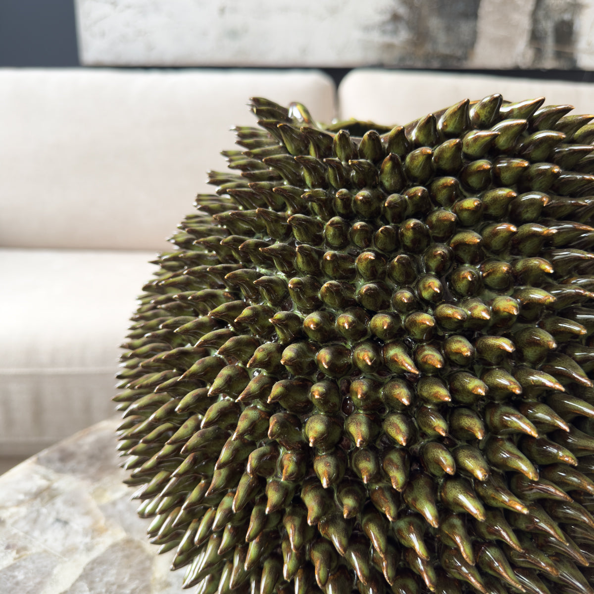 A green urchin vase, covered in 3D spikes, sitting infront of a cream sofa.