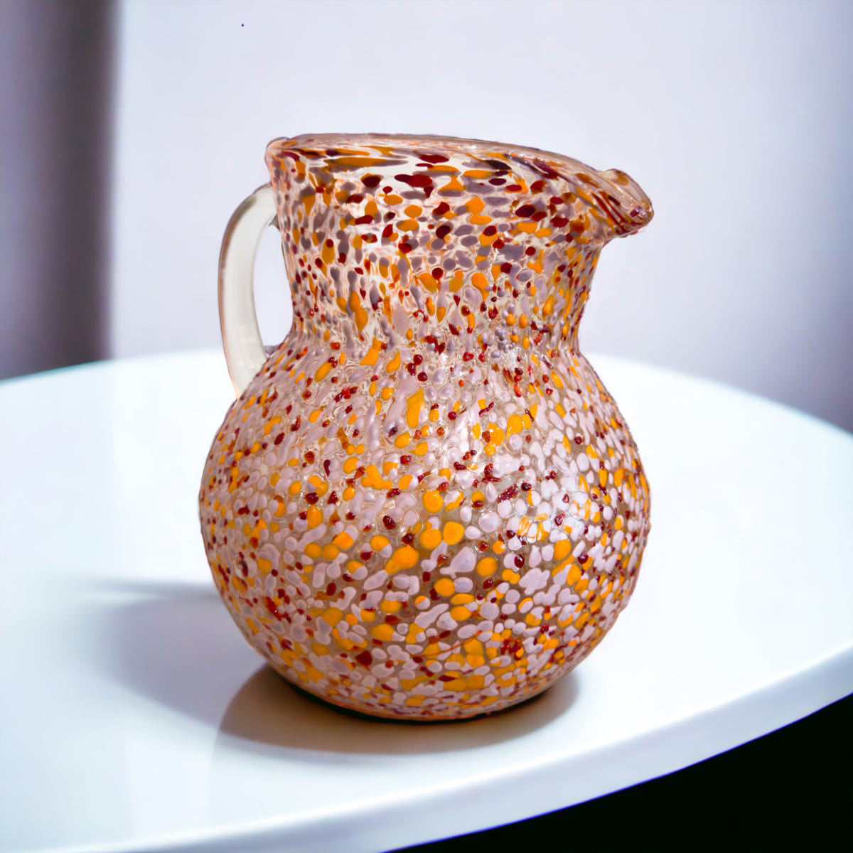 A multicolored glass jug with a confetti pattern, featuring shades of pink and orange, displayed on a white surface.