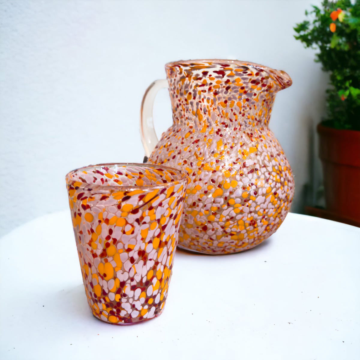 Speckled glass pitcher and cup on a white surface with a blurred plant in the background
