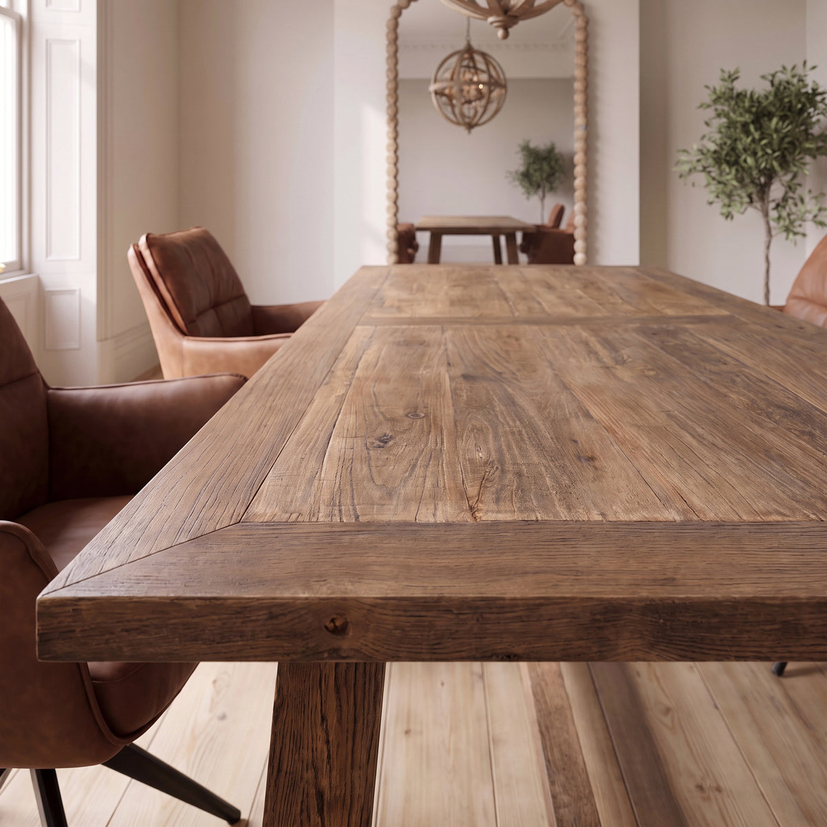 A rustic rectangular wooden dining table with a thick reinforced apron and visible wood grain, shown in a bright dining room with brown leather chairs and a mirror in the background.