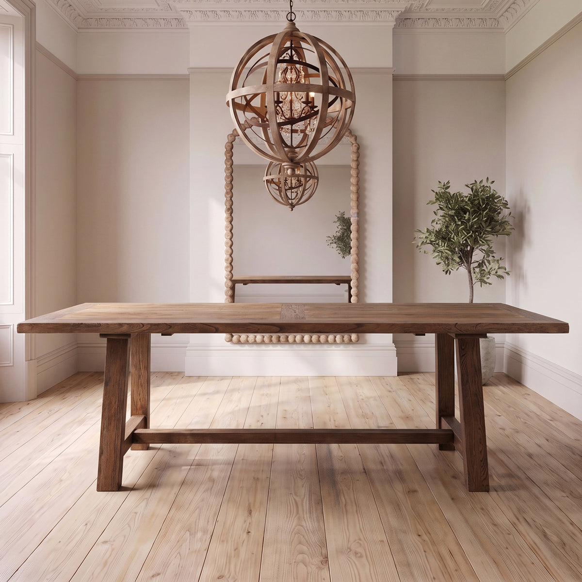 A wooden dining table with thick rectangular tops and trestle-style legs, shown in a bright, neutral-toned room with a large mirror, hanging globe chandeliers, and a potted plant.