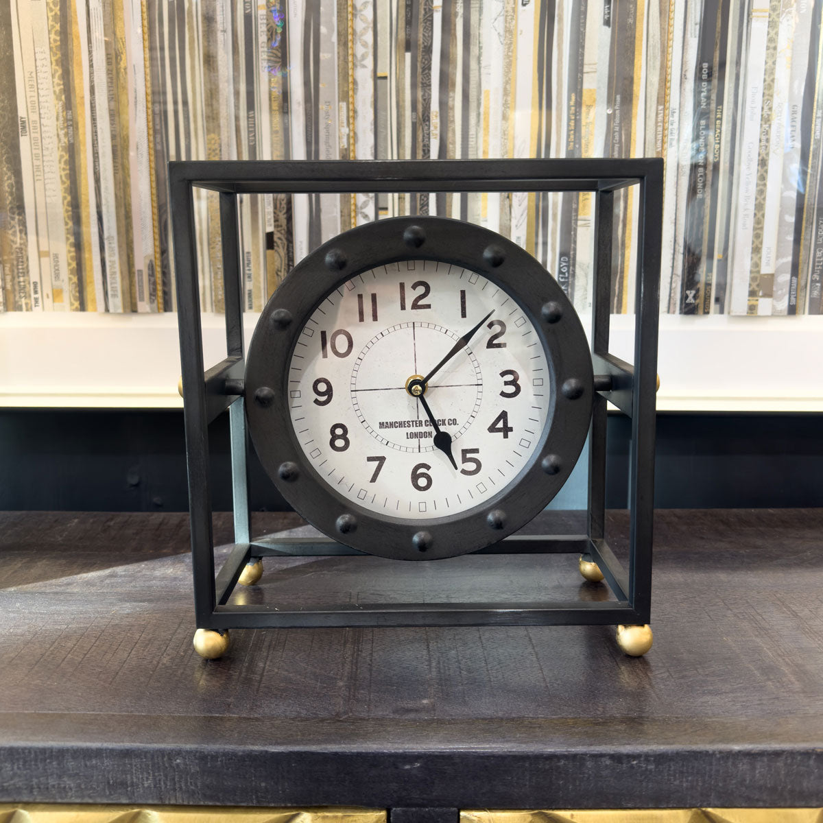 Decorative square black clock with black frame and white face on a wooden surface.
