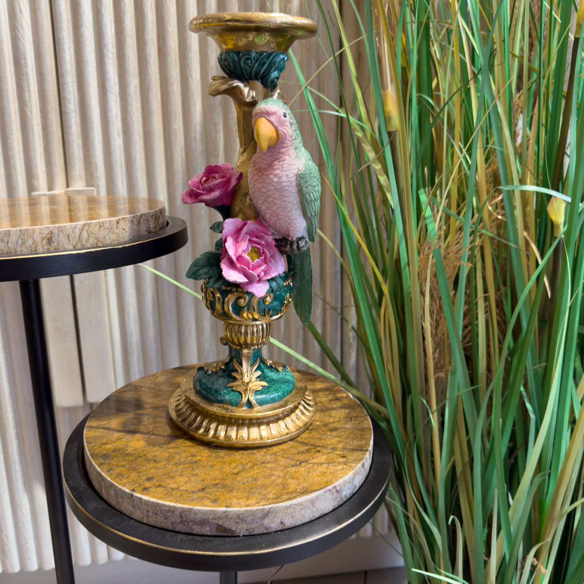 Ornate vintage parrot candlestick with pink flowers stands on a marble-topped table, framed by tall green grasses in an elegant room.