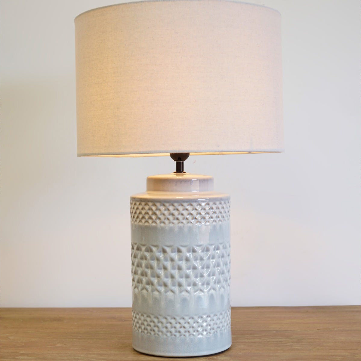 A lit pale blue table lamp with geometric pattern, sitting on a wooden table.
