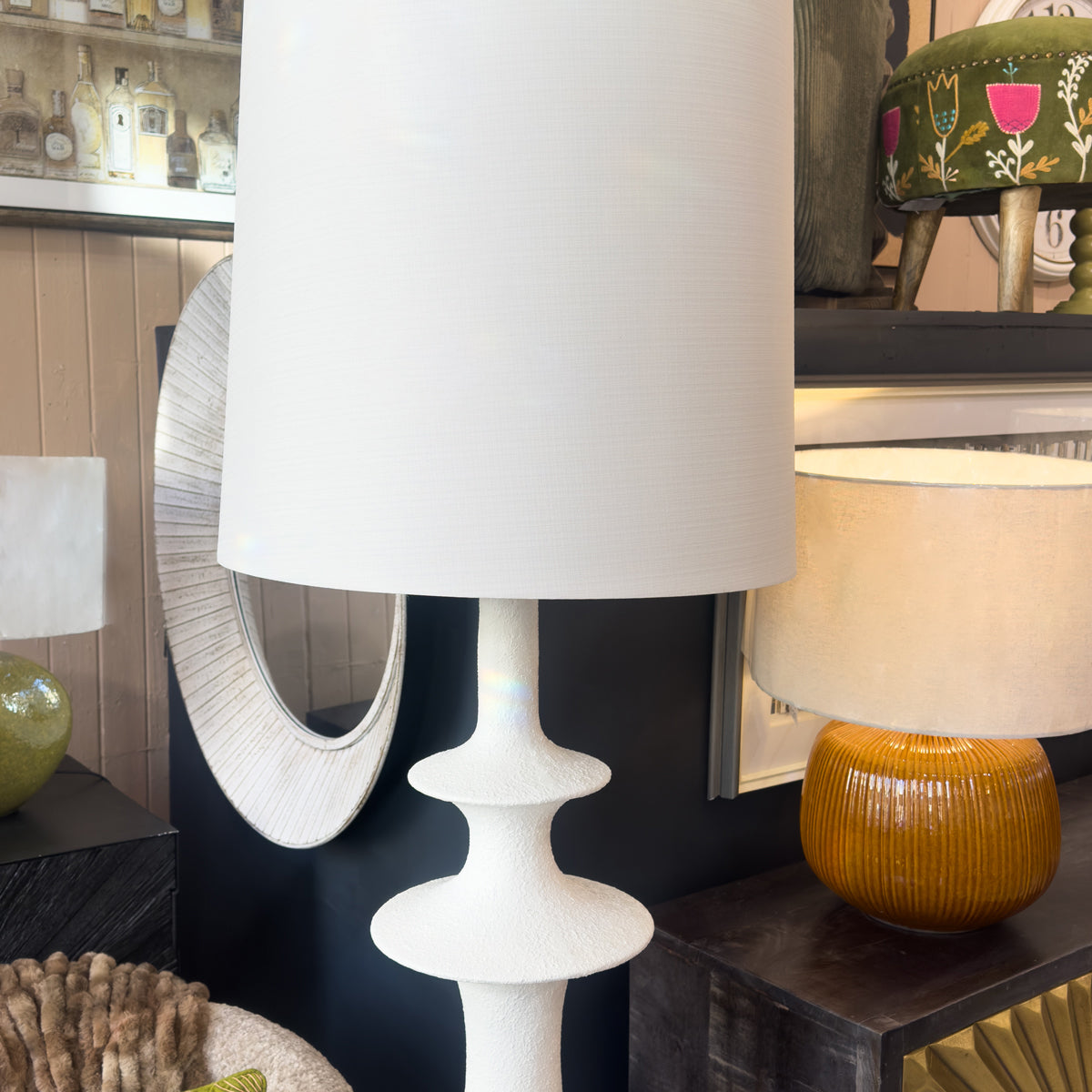White lamp with a textured base on a dark surface, with another lamp and decorative items in the background.