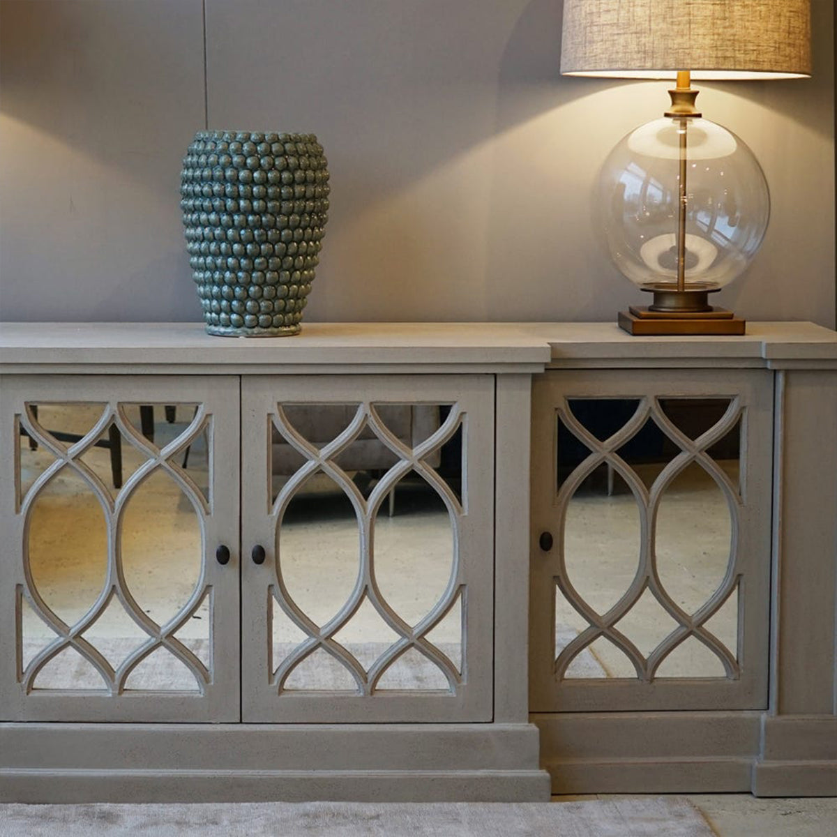 Whitewashed mirrored sideboard with decorative glass doors, a vase, and a lamp on top.