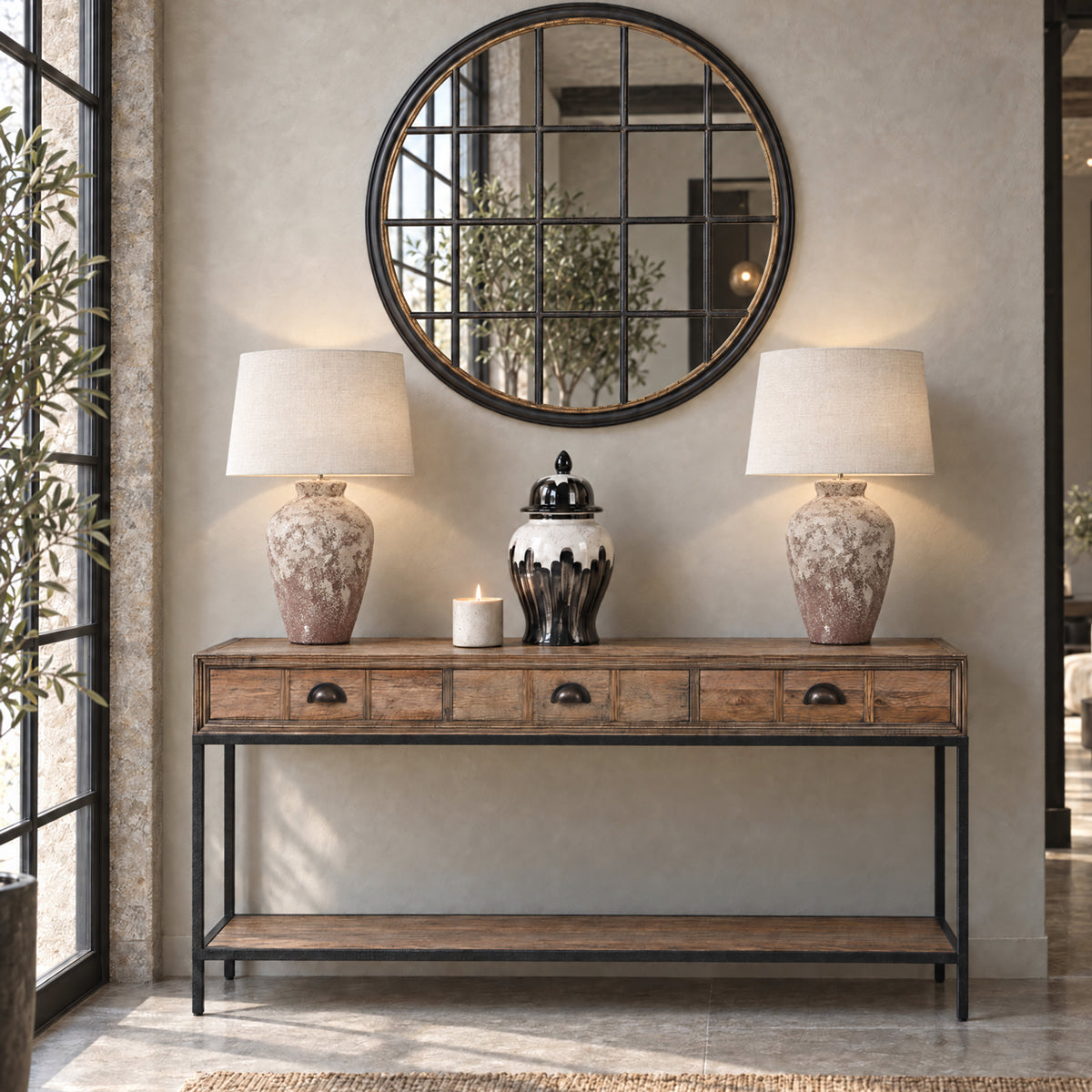 A rustic wooden console table with a black metal frame and lower shelf, styled in a home entryway with two matching table lamps, a white candle, and a round black-framed mirror on the wall.