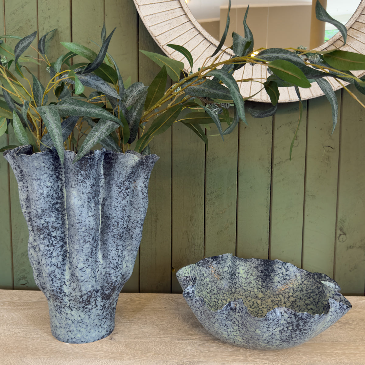 Decorative blue fluted speckled ceramic vase with leafy branches beside a matching shallow bowl, set against green wood paneling.