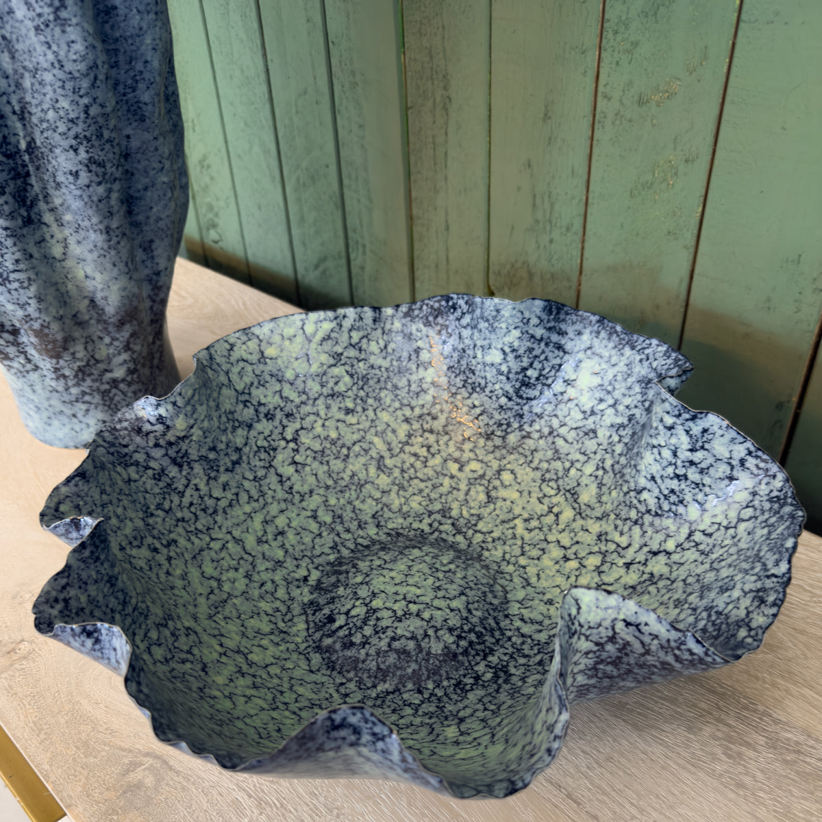 Large fluted ceramic bowl with blue speckled glaze sits on a wooden table against a weathered green plank wall, creating a rustic mood.