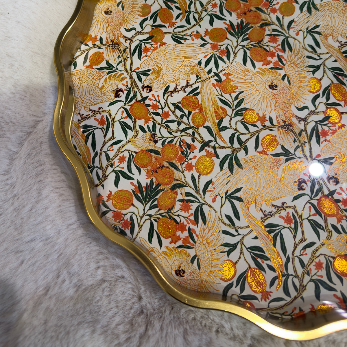 Ornate gold-edged tray with cream birds, leafy branches, and orange fruit motifs on a gray fur surface; decorative, warm, luxurious tone.