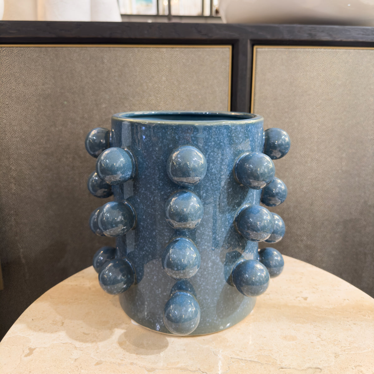 A blue plant pot with bobble details around the outside, sitting on a cream side table infront of a shagreen sideboard.