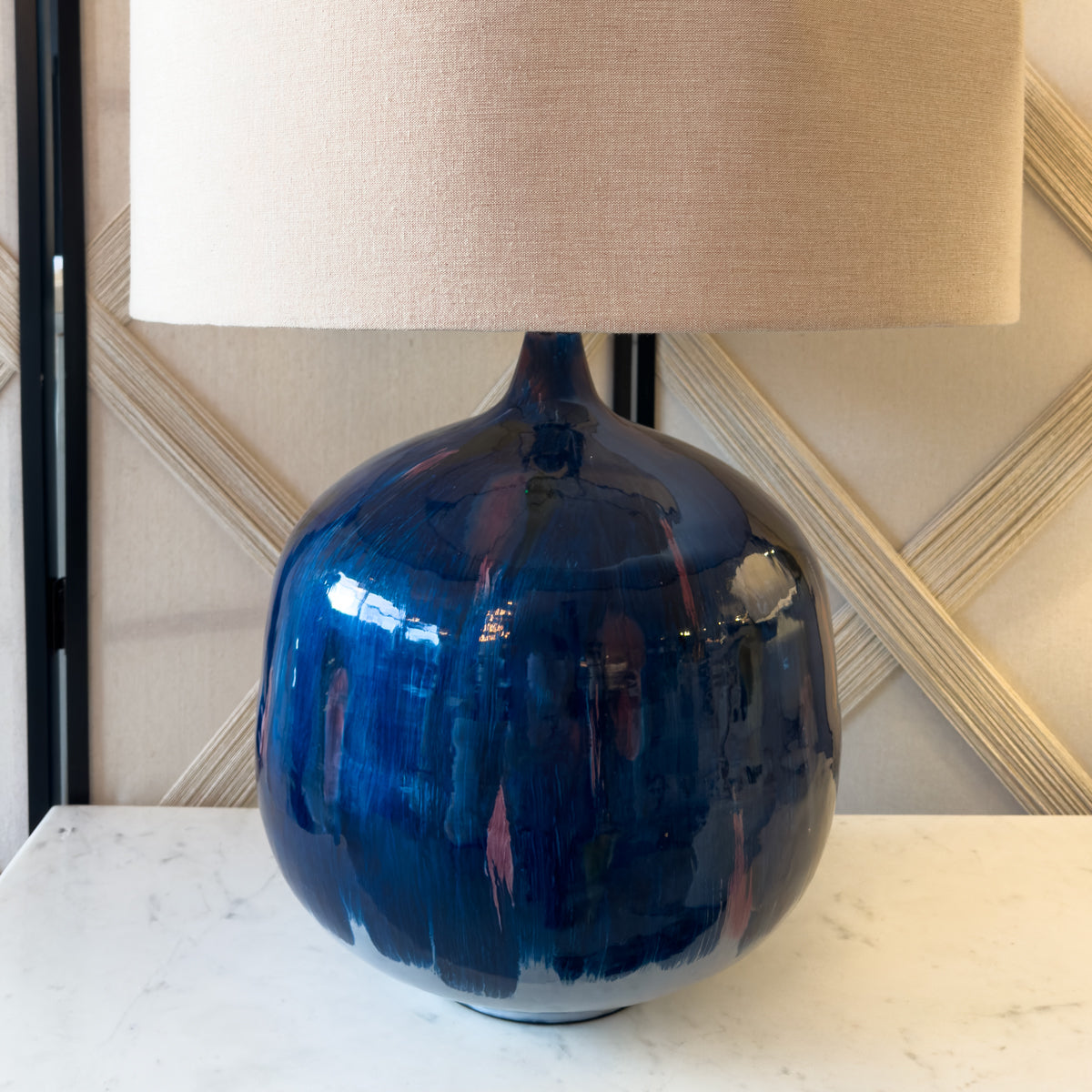 A blue and purple table lamp with a beige linen shade sitting on a white table.