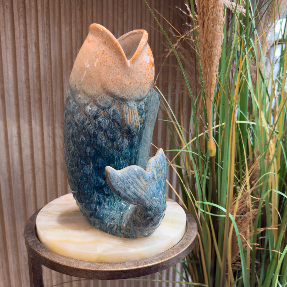 A blue and peach fish-shaped sculpture on a round marble pedestal, set against a ribbed wooden wall with tall grasses beside it.