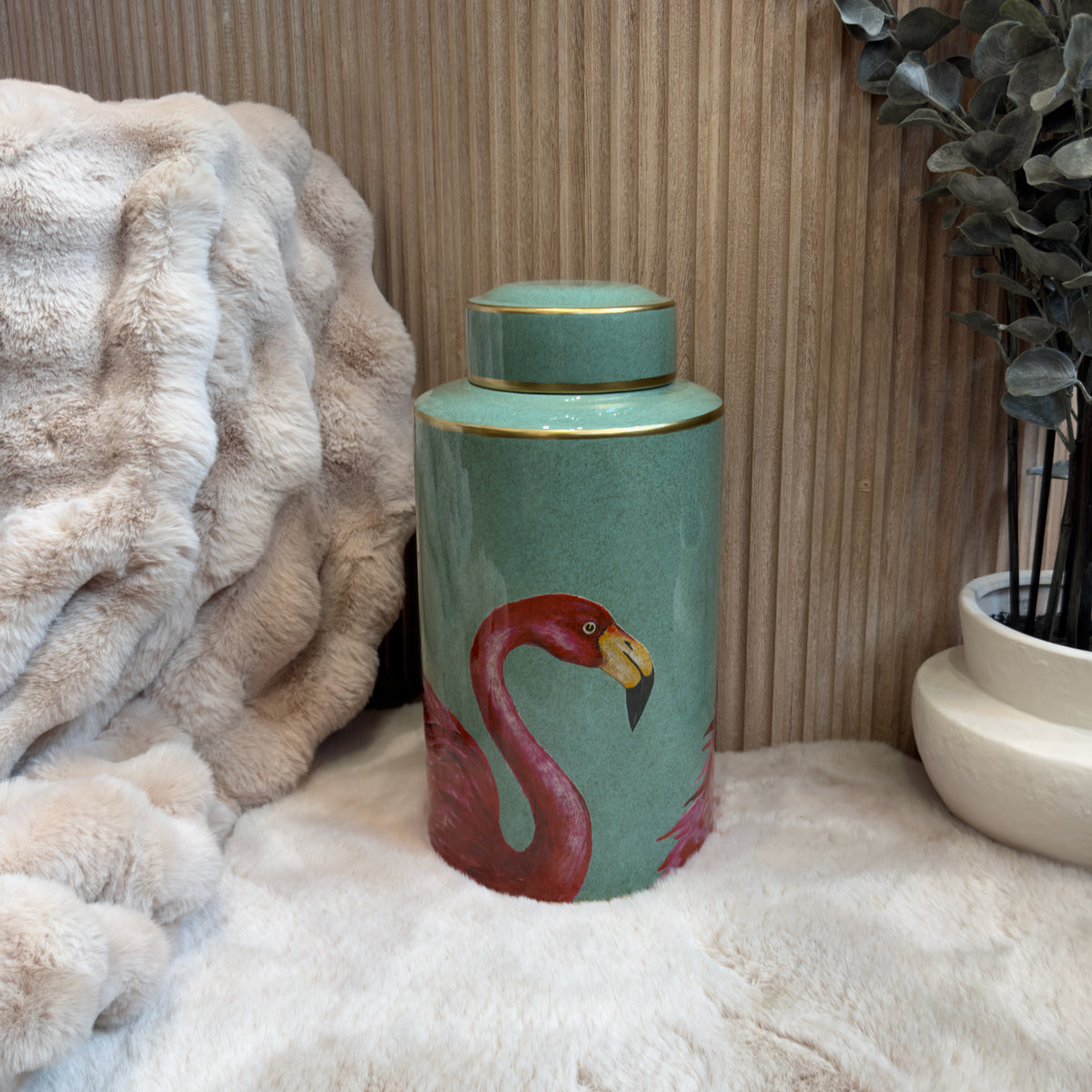 A cylindrical turquoise jar with a gold lid, featuring a painted pink flamingo on its side. The jar is placed on a soft beige fur surface adjacent to a potted plant and a textured backdrop of vertical lines.