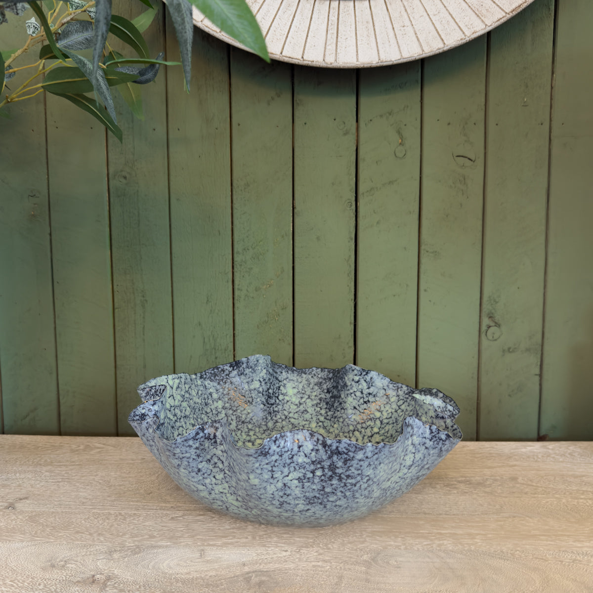 Fluted speckled glazed blue ceramic bowl sits on a wooden table against a green paneled wall, with leafy plants.