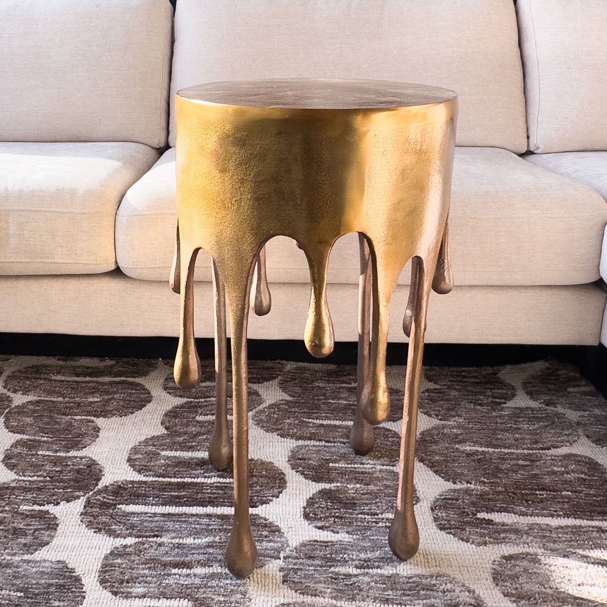 Gold side table with a drip design in front of a beige sofa on a patterned rug.