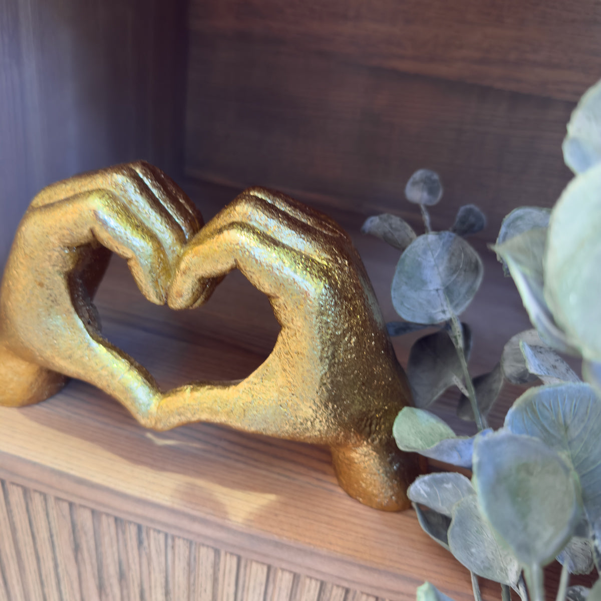 A gold sculpture of two hands forming a heart sits on a wooden shelf beside grey-green eucalyptus leaves.
