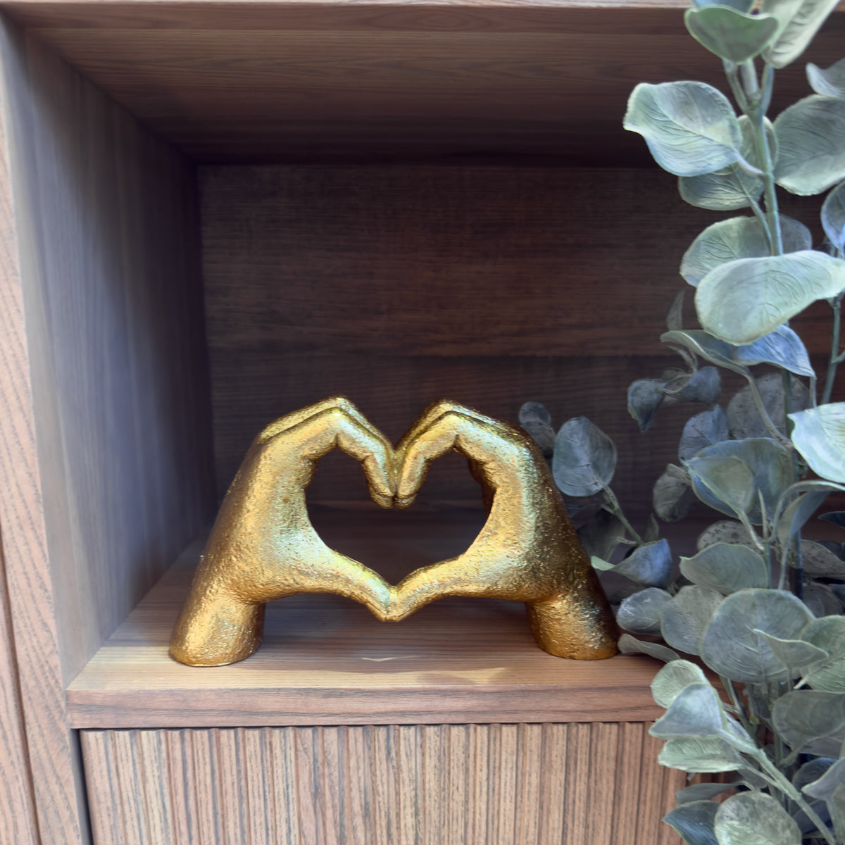 A gold sculpture of two hands forming a heart sits on a wooden shelf beside grey-green eucalyptus leaves.