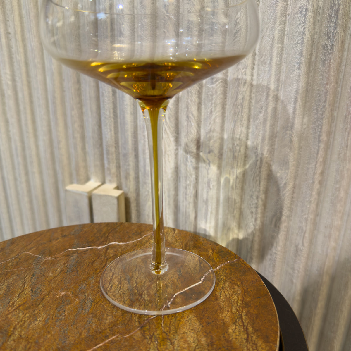 One glass clear cocktail glasses with golden stem stand on a round marbled table against pale textured sideboard.