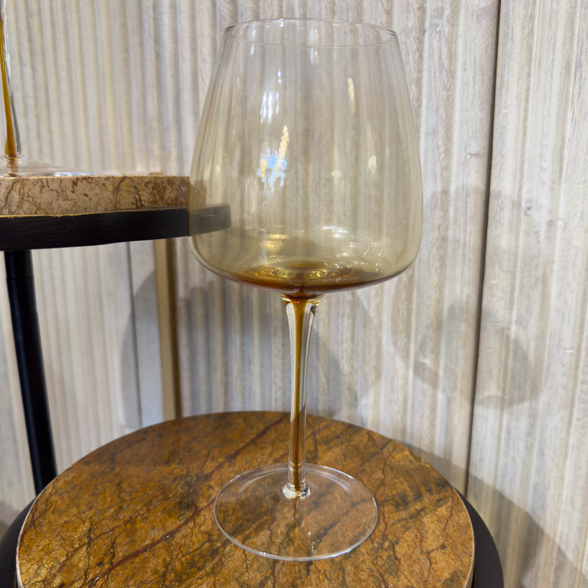 One glass wine glass with golden flooded detail stand on a round brown marble table beside a washed wood sideboard. 
