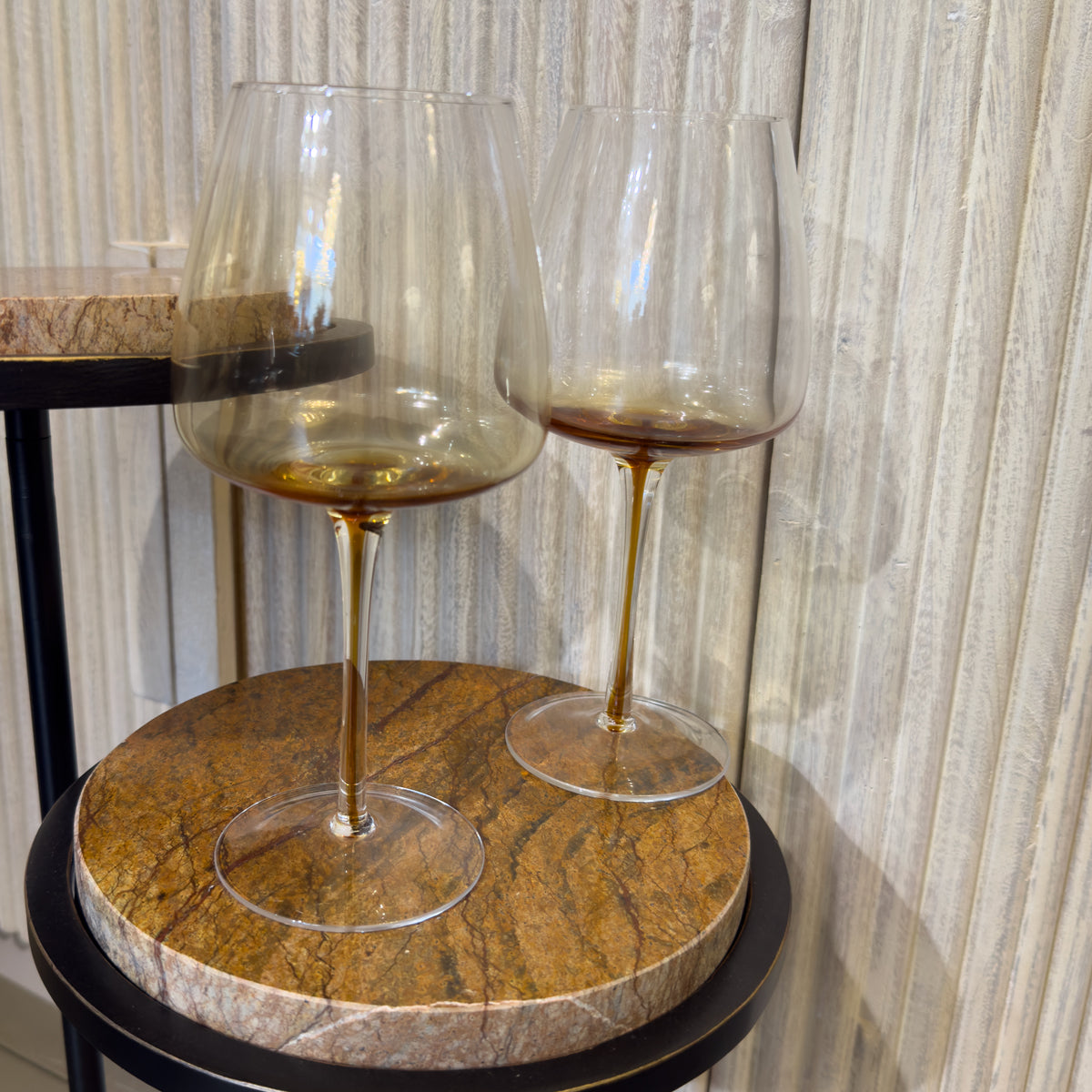 Two glass wine glasses with golden flooded detail stand on a round brown marble table beside a washed wood sideboard. 