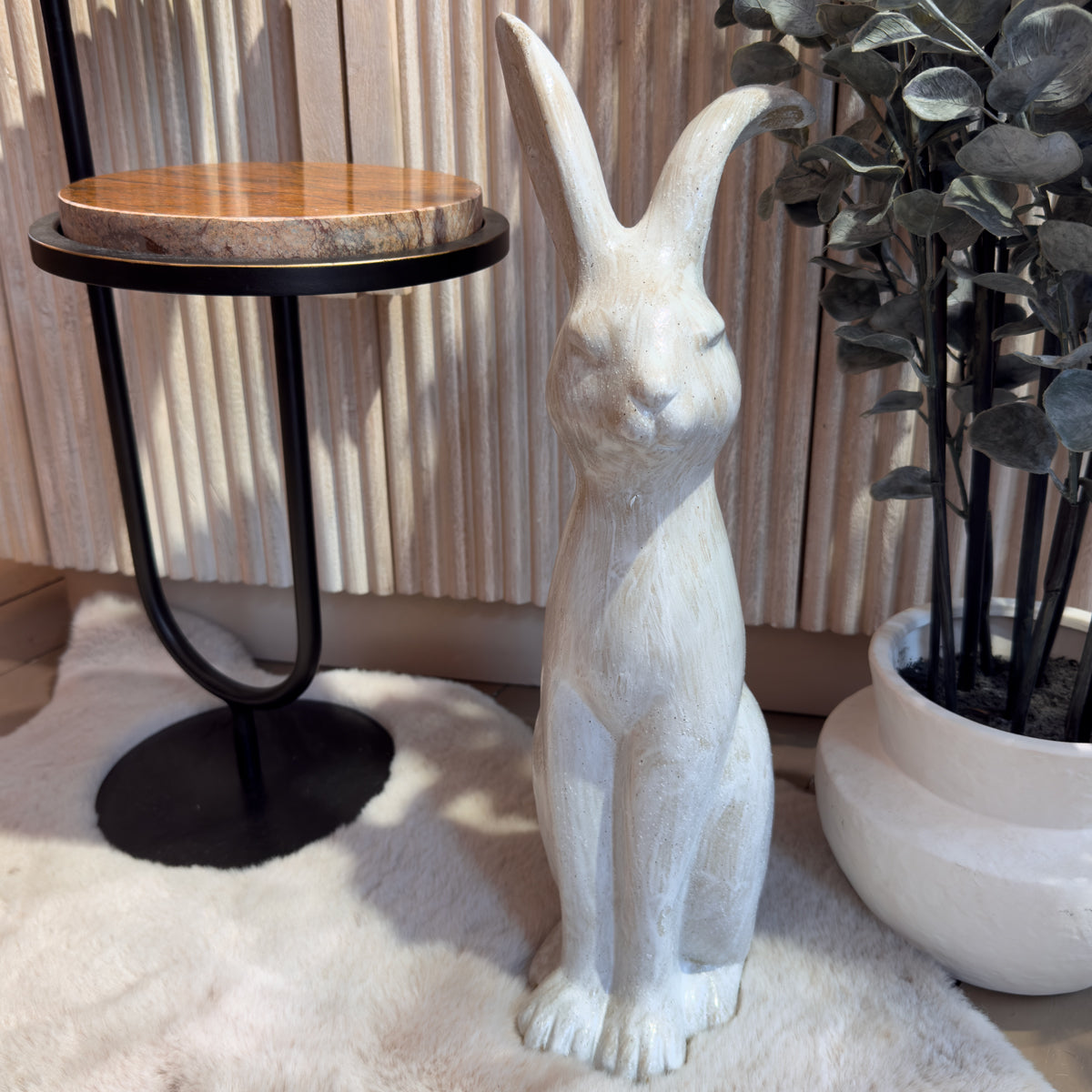 White rabbit statue stands beside a round wooden side table and eucalyptus potted plant, creating a serene, cozy interior scene with soft sunlight.
