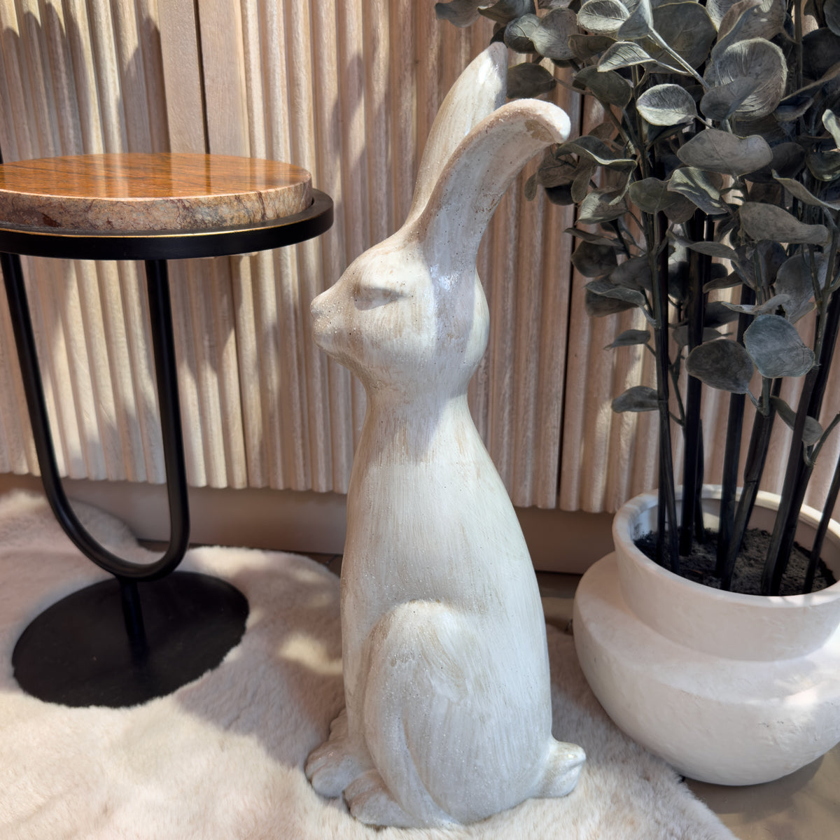 Side view of a white rabbit sculpture sits beside a round wood-and-metal table and potted eucalyptus plant, creating a warm, serene interior scene.