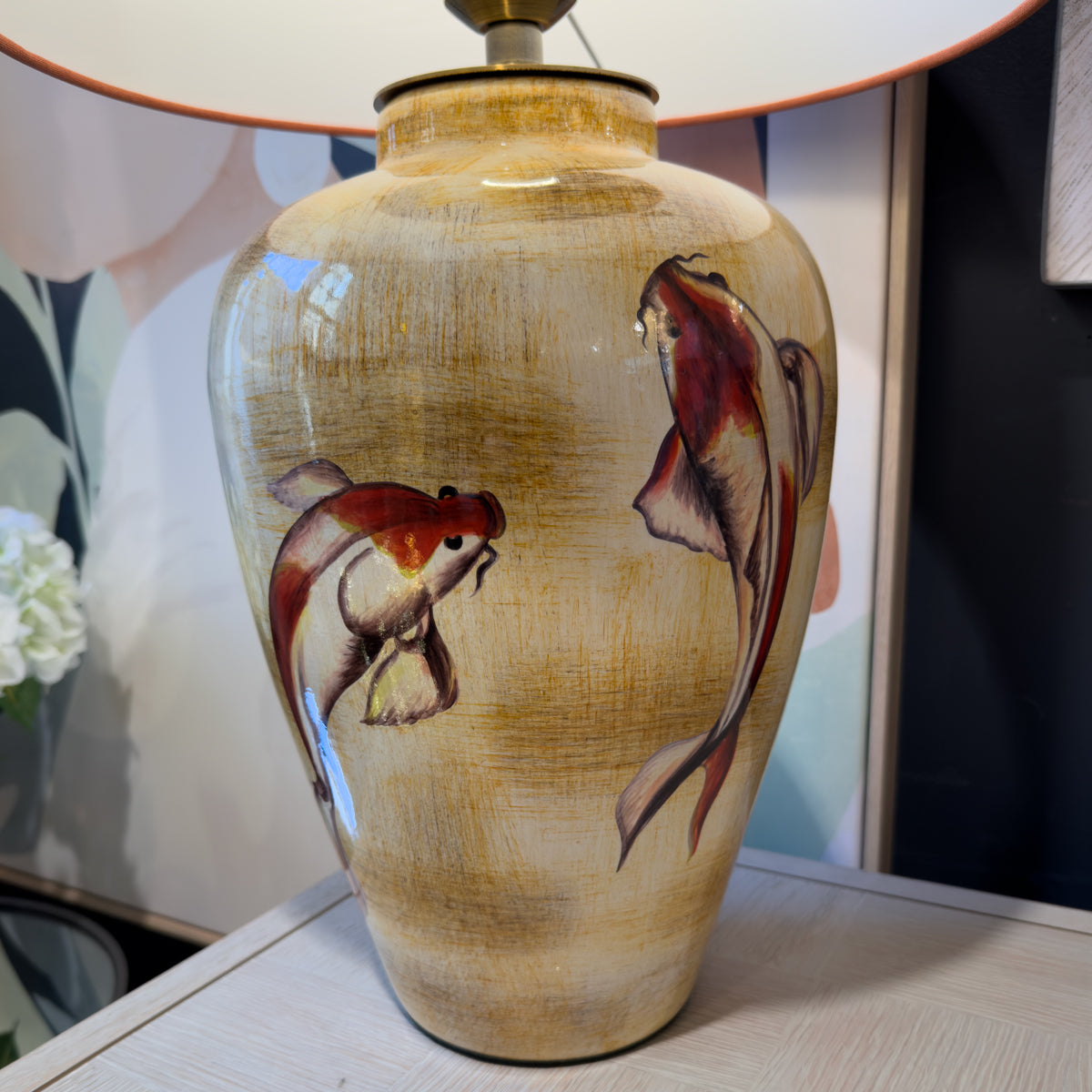 A ceramic table lamp base with a glossy beige and brown textured finish, decorated with two painted koi fish in red, orange, and dark brown. The lamp is shown sitting on a light-colored wooden table with a blurred background.