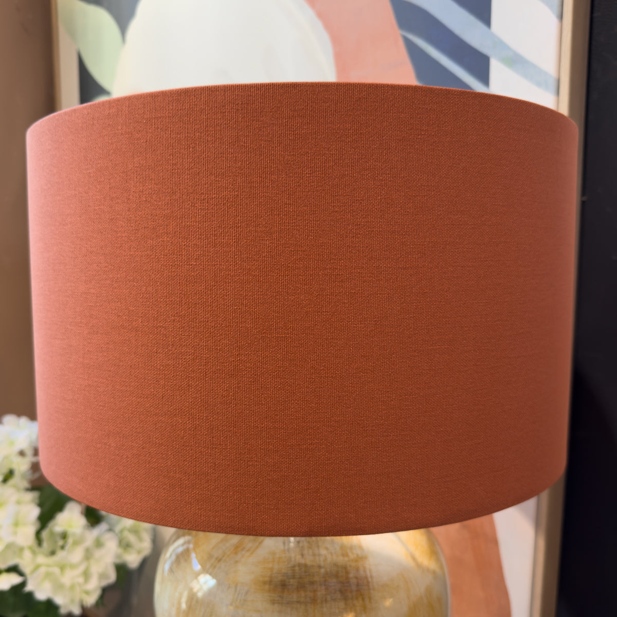 A close-up of a cylindrical fabric lampshade in a warm  rust orange color, mounted on a ceramic table lamp base.