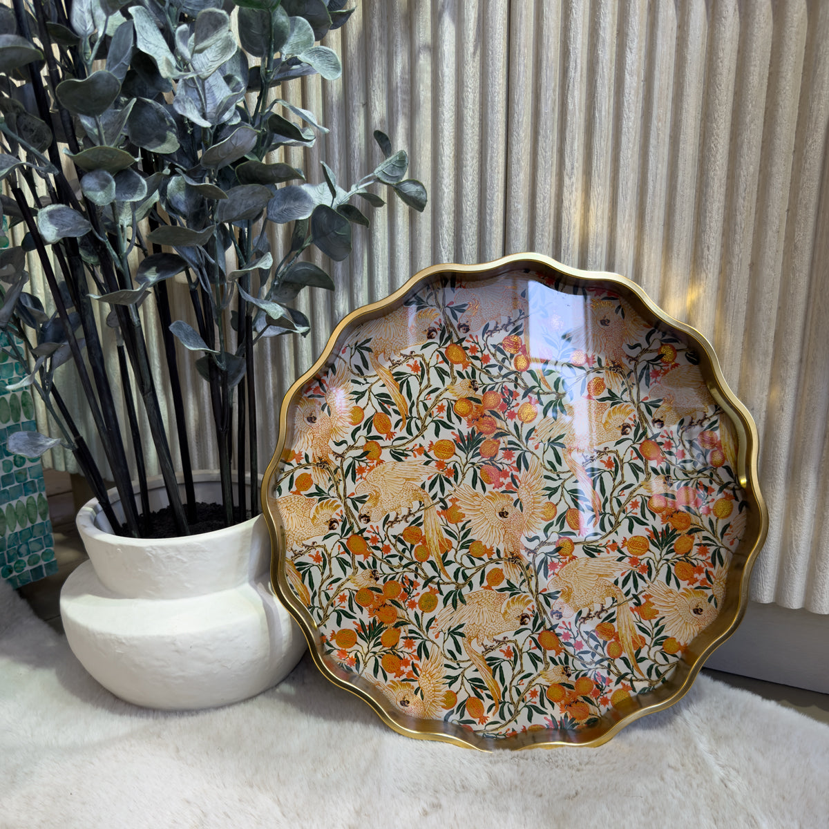 Scalloped gold-edged tray with colorful floral and cockatoo motifs beside a leafy potted plant, creating a warm, elegant interior scene.