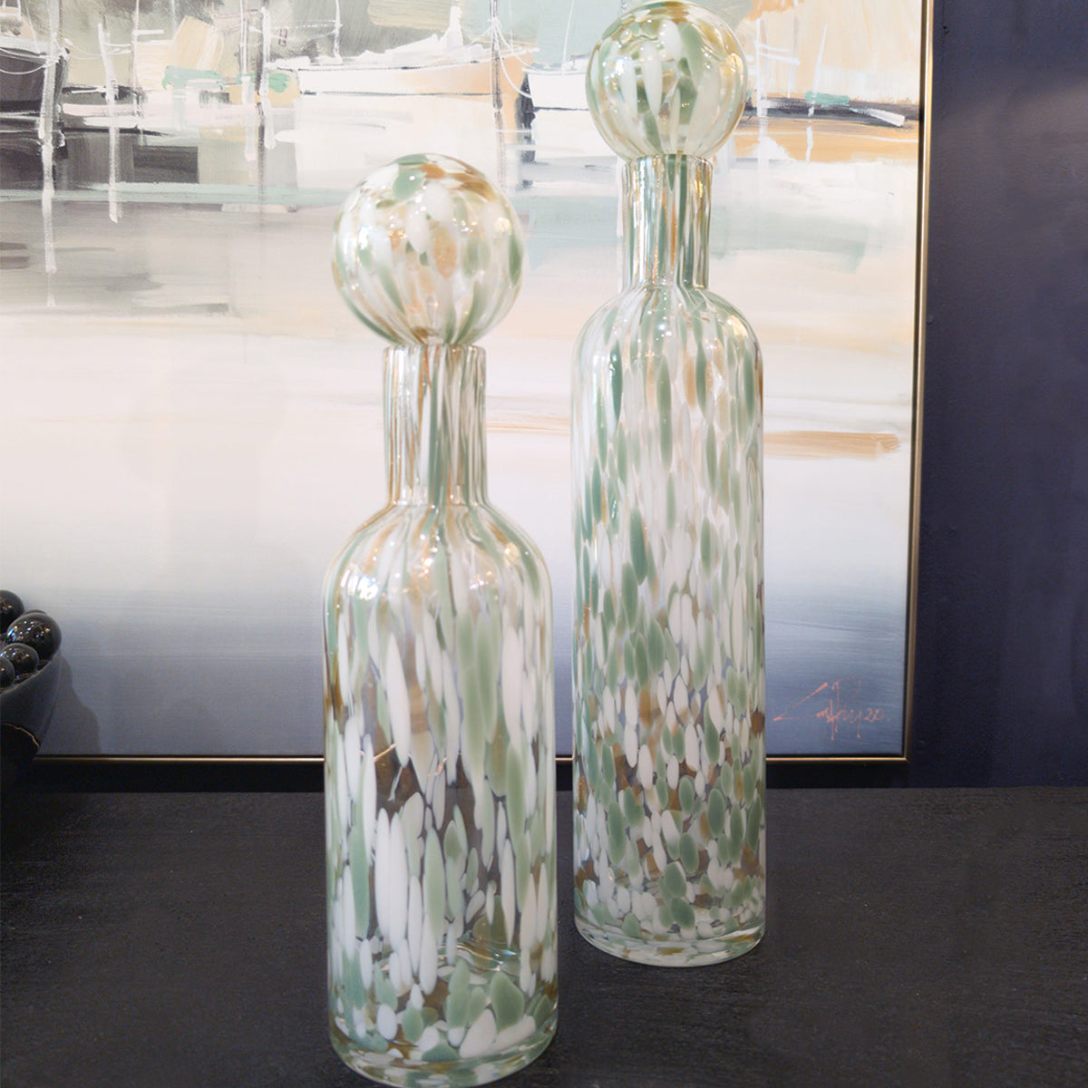 A set of sage green speckled vases with spherical lids, sitting on a black table infront of a coastal inspired canvas.