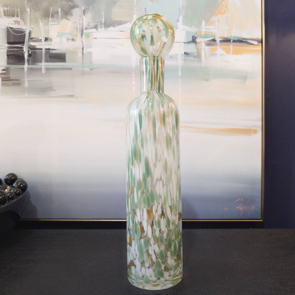 A tall and narrow green glass vase sitting on a black surface infront of a coastal canvas featuring docked boats.